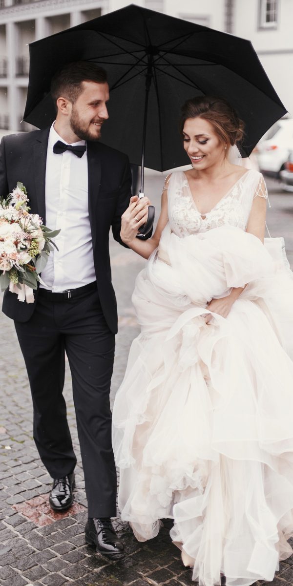 gorgeous-bride-and-stylish-groom-walking-under-umbrella-in-rainy-street-and-smiling.jpg
